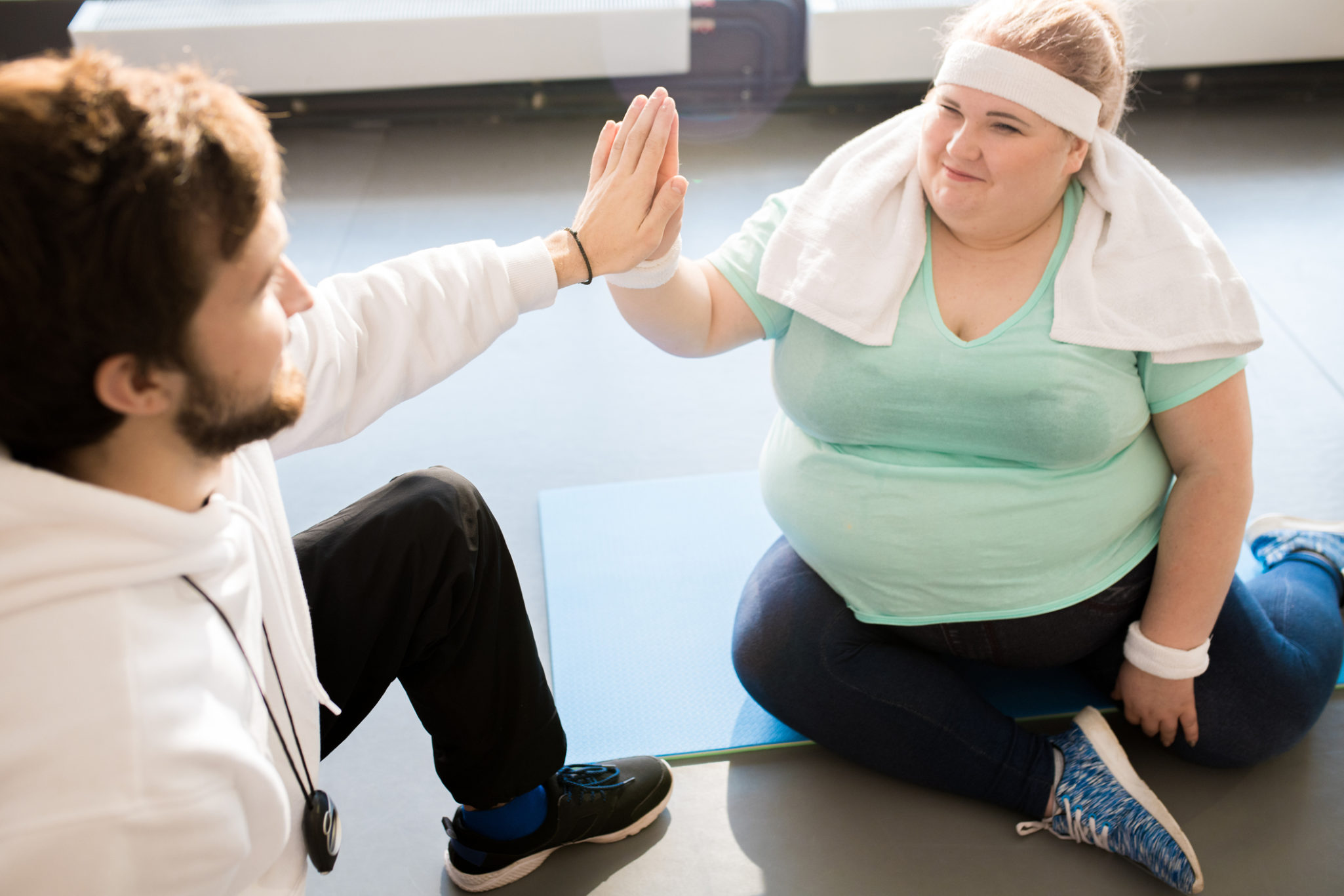 High angle portrait of smiling obese young woman sitting on floor and doing high five with coach while taking break in workout
